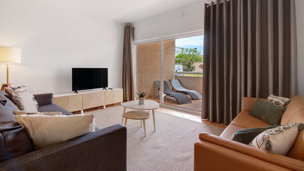 A modern hotel room with a sofa, armchair, TV, and a balcony with a view.