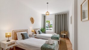 A hotel room with two beds, a window with curtains, a chair, and a hanging light fixture.