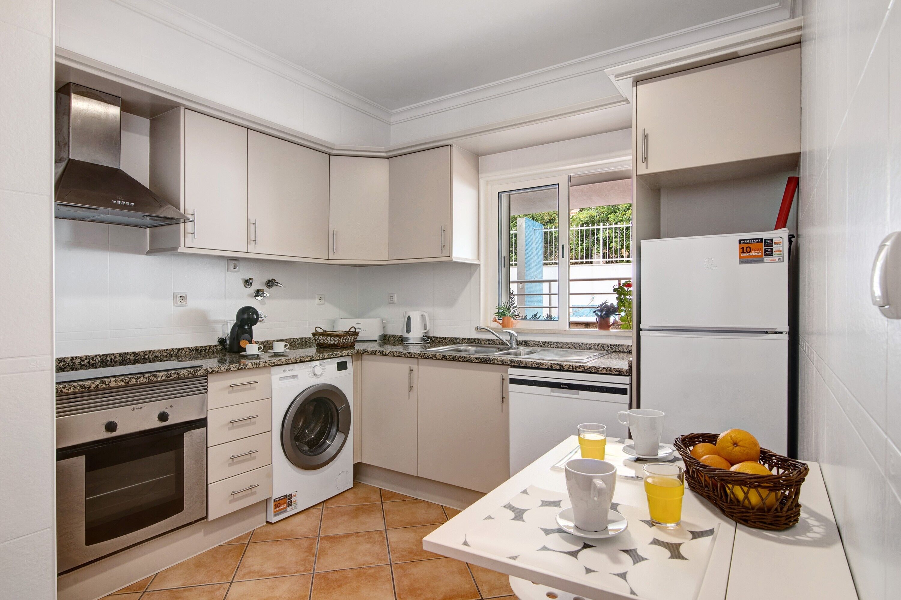 Une cuisine équipée d’un lave-linge, d’un four et d’un réfrigérateur, une table à manger avec un panier de fruits, et une fenêtre donnant sur un paysage verdoyant.