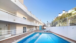 Une piscine avec une balustrade blanche, une terrasse ensoleillée et une vue sur un immeuble d’habitation.