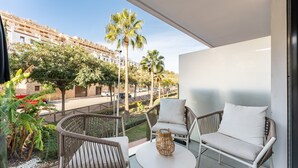 A balcony with wicker furniture, a small table, and a view of a street with palm trees and buildings.