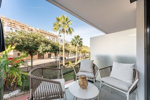 A balcony with wicker furniture, a small table, and a view of a street with palm trees and buildings.