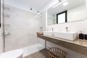 A modern bathroom with a glass shower enclosure, two sinks, a large mirror, and a wicker chair.