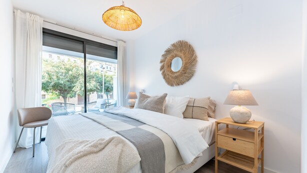 A bedroom with a bed, a nightstand, a lamp, a chair, a window with curtains, and a wall decoration.