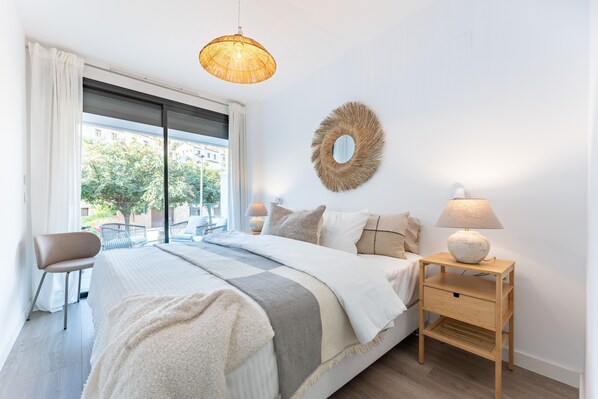 A bedroom with a bed, a nightstand, a lamp, a chair, a window with curtains, and a wall decoration.