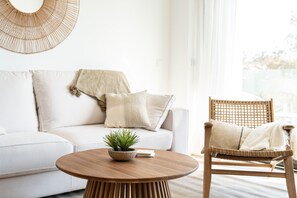 A modern living room with a white sofa, a wooden chair, a round wooden coffee table, and a woven wall decoration.