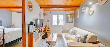Ein gemütliches Wohnzimmer mit Holzbalkendecke, einer Couch, einem Fernseher, einer Gitarre und einem Fenster.