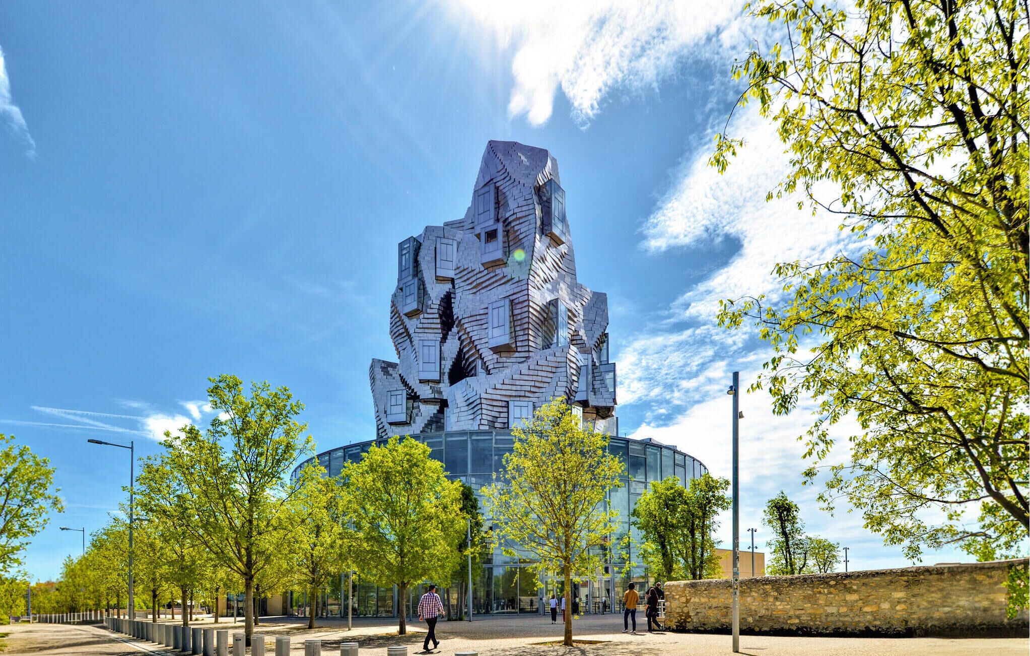 A modern building with a unique, angular design, surrounded by a park with trees and a clear sky.