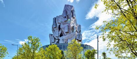 A modern building with a unique, angular design, surrounded by a park with trees and a clear sky.