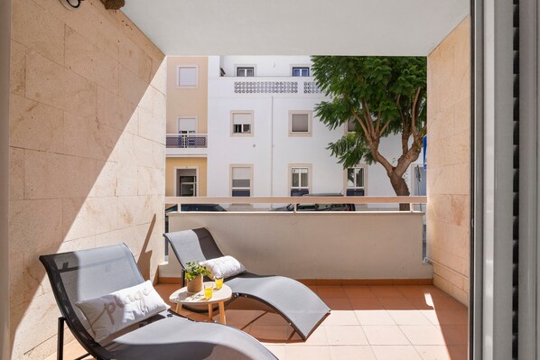 Un balcon avec deux fauteuils, une petite table ornée d’une plante et d’un verre de jus d’orange, et une vue sur un immeuble doté de balcons et sur un arbre.