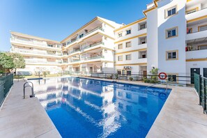 Una piscina con superficie de agua azul cristalina, rodeada por un edificio de varios pisos con balcones y fachada blanca.