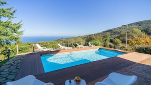 A swimming pool with lounge chairs and a table with fruit, surrounded by a wooden deck and stone pathway, overlooking a scenic hillside and ocean. - Awesome home in Caronia (Caronia)
