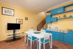 Un comedor con televisión, sillas de mimbre blancas, una mesa y una cocina con armarios azules.