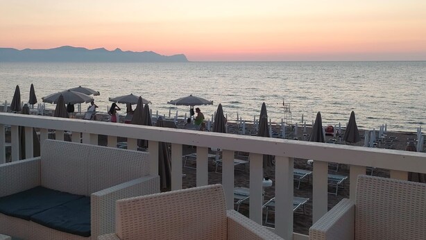 Une terrasse en bord de mer, dotée de meubles en osier, de parasols et offrant une vue sur l’océan et les montagnes lointaines.