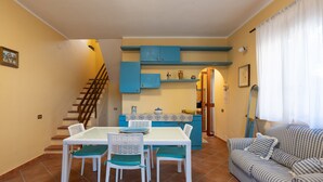 Un coin repas avec une table et des chaises blanches, une cuisine avec des placards bleus, un escalier et un canapé à rayures.