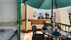 En balkon med en seng, et bord med stole, en grøn parasol og en pool i baggrunden.
