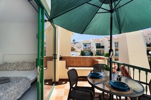 A balcony with a bed, a table with chairs, a green umbrella, and a pool in the background. - Vacation Apartment 'Mareverde K131' with Shared Pool, Balcony and Wi-Fi (Adeje)