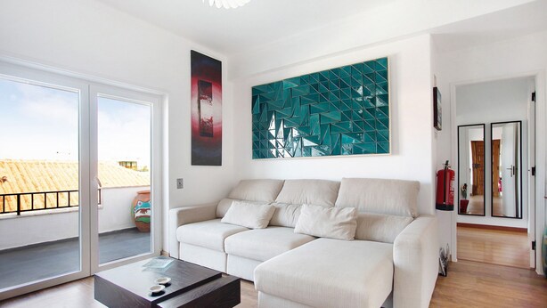 A living room with a sofa, a coffee table, a large window, and a geometric wall art piece. - Apartamento Da Alegria – Feel at Home on the Silver Coast (Caldas da Rainha)