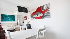 A modern living room with a white sofa, a dining table with chairs, and a large abstract painting on the wall. - Apartamento Da Alegria – Feel at Home on the Silver Coast (Caldas da Rainha)