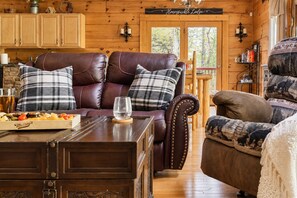 A cozy cabin interior with a leather sofa, plaid cushions, a wooden coffee table, and a dining table with a fruit platter. - Free Dollywood Tix/Dog Friendly/Gameroom/HotTub/ (Sevierville)