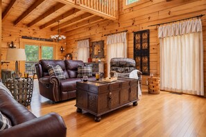 A cozy cabin interior with a wooden ceiling, a leather sofa, a wooden coffee table, and a large window with curtains. - Free Dollywood Tix/Dog Friendly/Gameroom/HotTub/ (Sevierville)