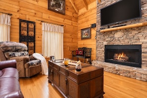A cozy log cabin interior with a fireplace, wooden walls, a leather couch, and a dining table with a glass of wine. - Free Dollywood Tix/Dog Friendly/Gameroom/HotTub/ (Sevierville)