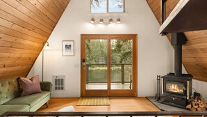 A cozy room with a green sofa, a fireplace, and a view of the outdoors through a glass door. - Stylish Lakefront Cabin Retreat, Sauna/Hot tub & Cold Plunge (Gig Harbor)