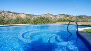 Ein klares, blaues Schwimmbecken mit einer Metallleiter, umgeben von einer Steinmauer und einer Rasenfläche, mit einer hügeligen Landschaft im Hintergrund.