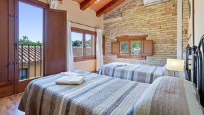 A bedroom with two beds, wooden ceiling beams, a stone wall, and a window with curtains.