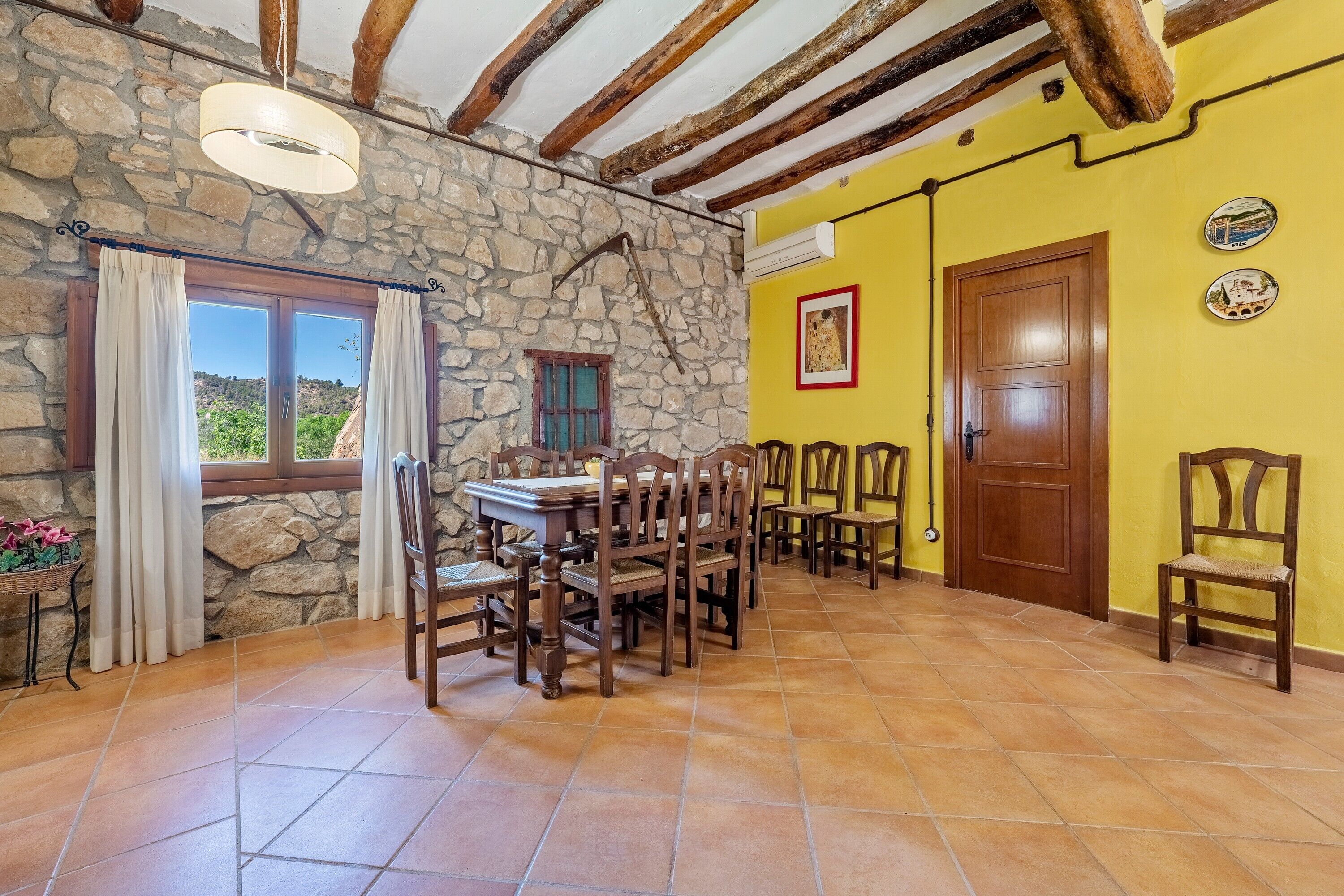 A dining area with a stone wall, wooden ceiling beams, a tiled floor, and a table set for four.