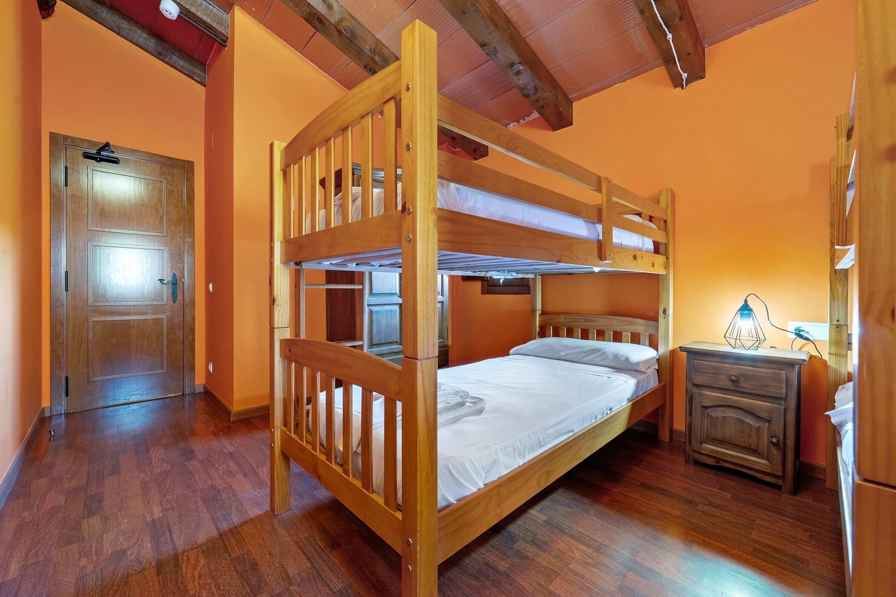 Une chambre avec un lit superposé, une porte en bois, une table de chevet avec une lampe et un plancher en bois.