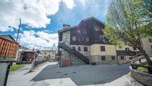 A traditional stone building with a staircase leading to the entrance, surrounded by a paved area and other buildings. - Comfortable and spacious apartment for four people (Livigno)