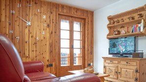 A living room with a wooden wall clock, a red leather sofa, a wooden cabinet with books and decorative items, and a television on a wooden stand. - Comfortable and spacious apartment for four people (Livigno)