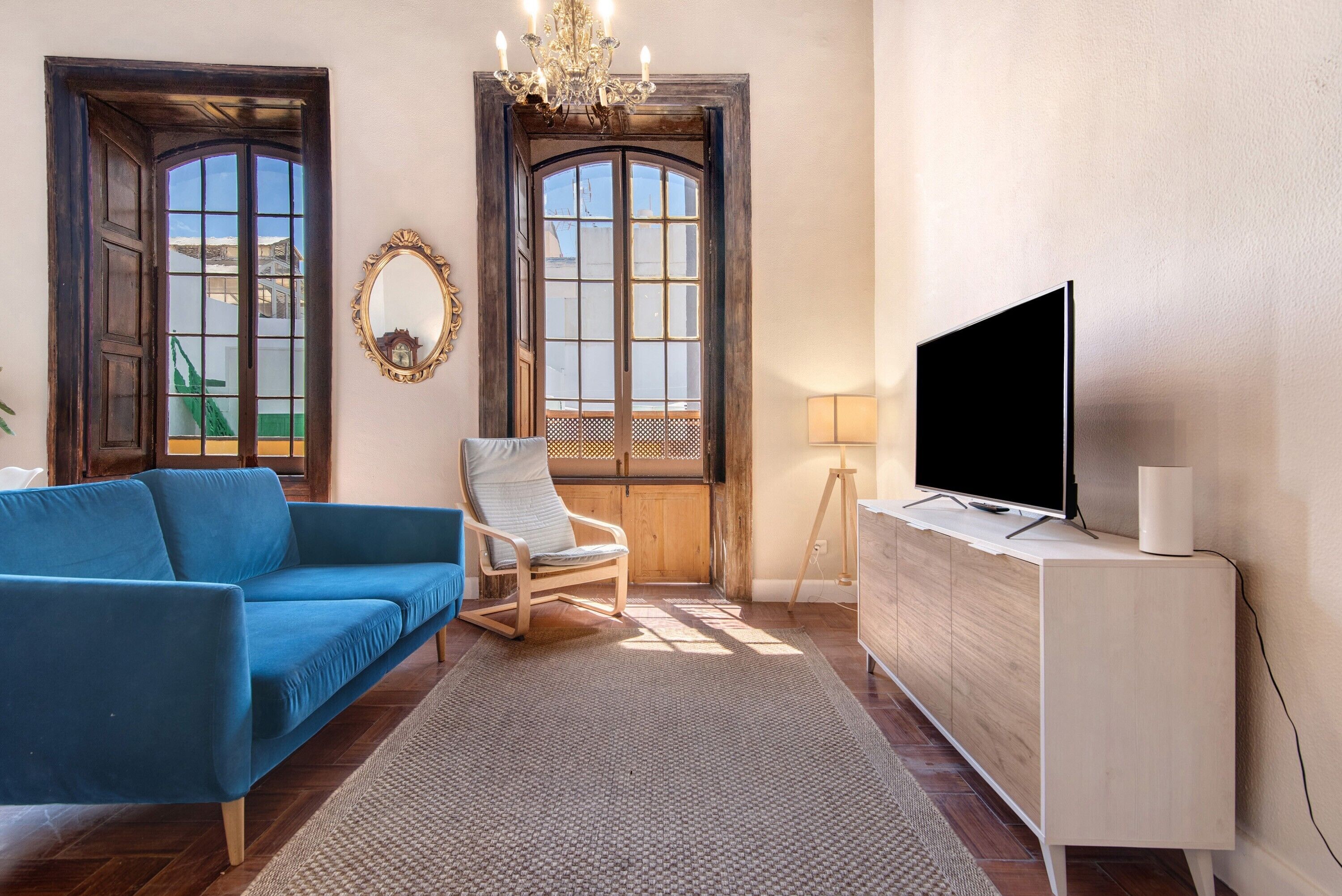 Una sala de estar con un sofá azul, una silla de madera, un televisor sobre un mueble bajo y un ventanal con vistas.
