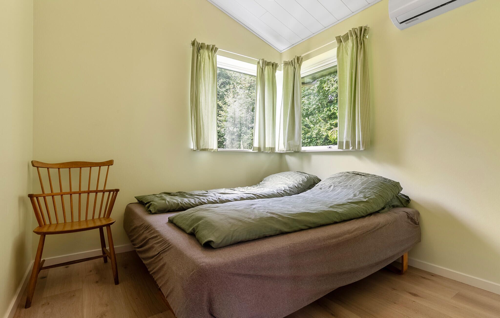 A small, single-bed room with a wooden chair, two windows with curtains, and an air conditioning unit.