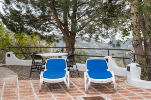 Una terraza con dos sillas azules y una mesa, rodeada de muros blancos y una barandilla, con vistas a un paisaje montañoso.