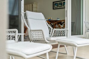 A set of white wicker lounge chairs with cushions, positioned on a balcony with a view of an indoor dining area.