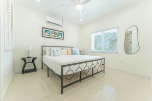 A bedroom with a bed, a bedside table, a ceiling fan, an air conditioning unit, a window with blinds, and a wall-mounted mirror.