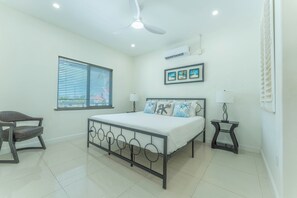 A bedroom with a bed, a chair, a bedside table, a lamp, a ceiling fan, and a window with blinds.