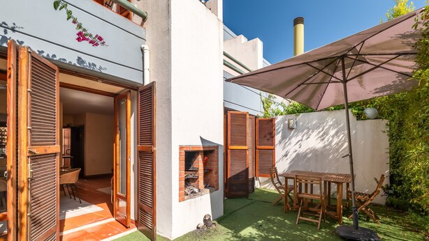 An outdoor patio area with a wooden table and chairs, a barbecue grill, and a large umbrella providing shade.