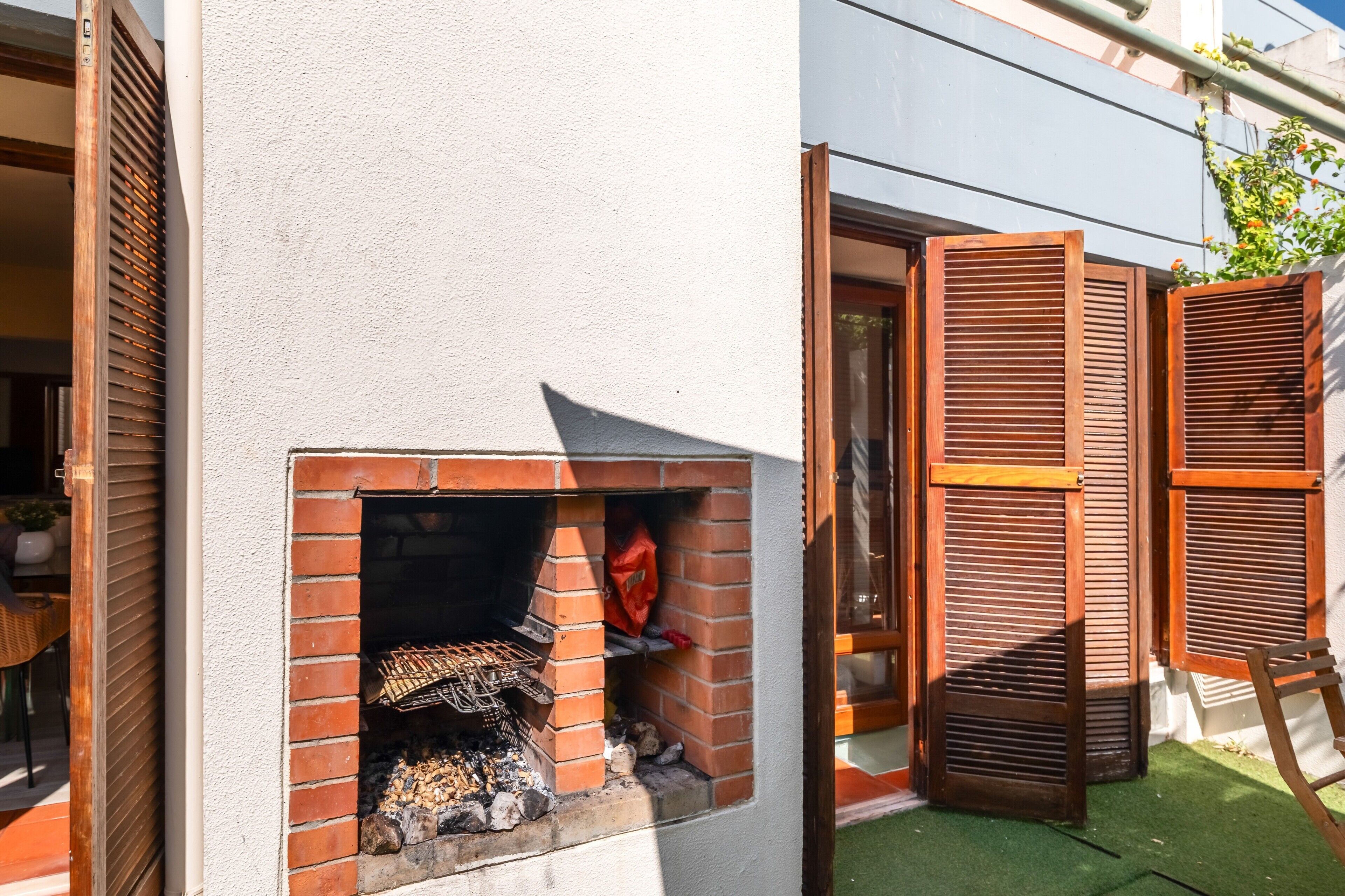 A brick-built outdoor grill area with a wooden shutter door, a small brick oven, and a wooden chair.