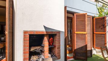 A brick-built outdoor grill area with a wooden shutter door, a small brick oven, and a wooden chair.