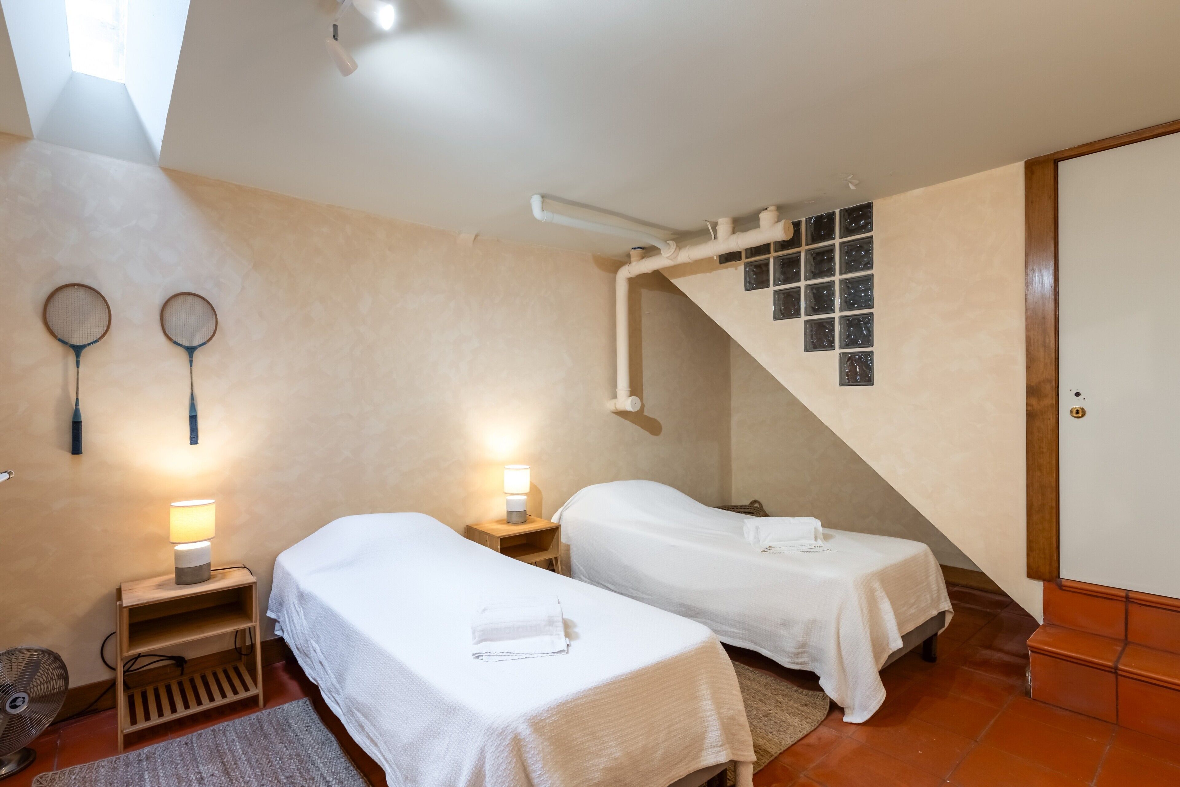 A bedroom with two single beds, a wooden nightstand with a lamp, a fan, and a window with blinds.