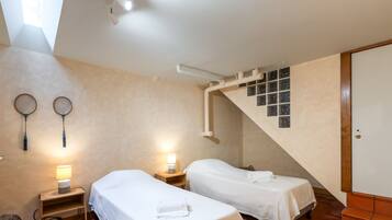 A bedroom with two single beds, a wooden nightstand with a lamp, a fan, and a window with blinds.
