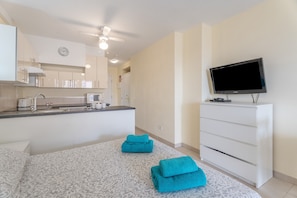 A modern hotel room with a kitchenette, a flat-screen TV, a bed with patterned bedding, and a ceiling fan.