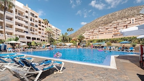 A swimming pool with lounge chairs, a mountainous backdrop, and a multi-story hotel building. - Vacation 'Castle Harbour' with Sea View, Shared Pool and Private Terrace (Los Cristianos)