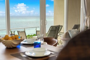 Un coin repas avec vue sur l’océan, une table sur laquelle se trouve un saladier rempli de fruits, une tasse et un verre contenant un liquide bleu.