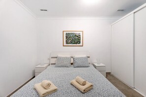 Een slaapkamer met een bed, twee kussens, een nachtkastje met een lamp, een ingelijste foto aan de muur en een handdoek op het bed.