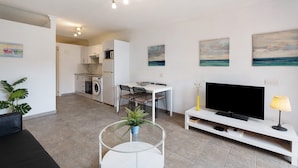 A modern living room with a flat-screen TV, a glass coffee table, a dining area with a white table and chairs, and a kitchen in the background. - Vacation Apartment 'Orlando 103' with Shared Pool, Private Terrace and Wi-Fi (Adeje)