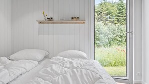 Ein Schlafzimmer mit einem Bett, weißer Bettwäsche, einer Holzablage mit Dekoration und einem großen Fenster mit Blick auf Grünfläche.